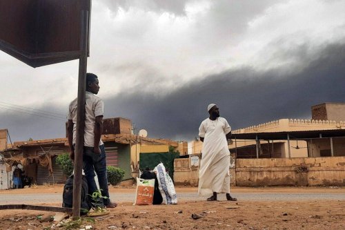 Smoke rises above buildings as people wait on the side of a road with some belongings, in Khartoum on June 10, 2023 [AFP via Getty Images]